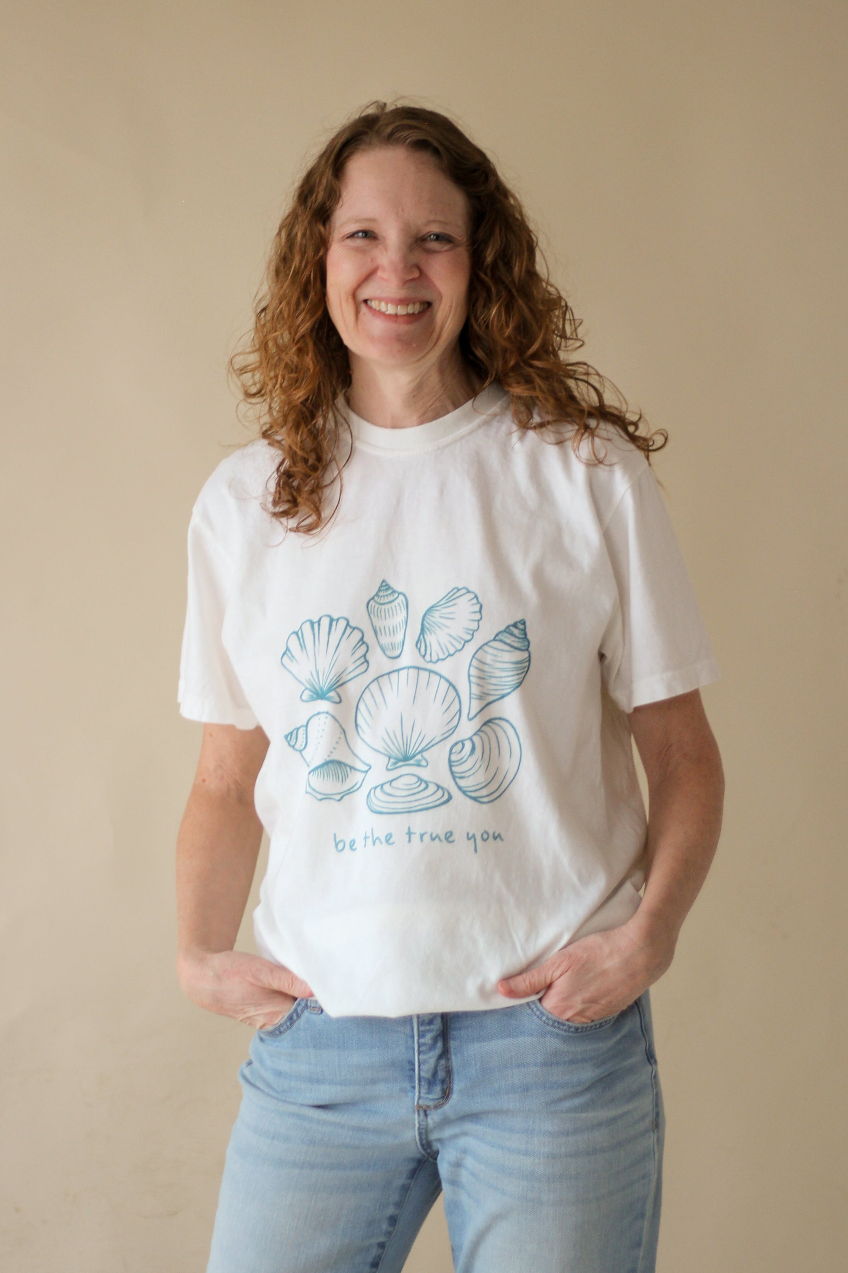 woman with medium length red curly hair wearing a white t-shirt on a beige background. The t-shirt features an illustrated printed graphic of an assortment of seashells with the text "be the true you" underneath