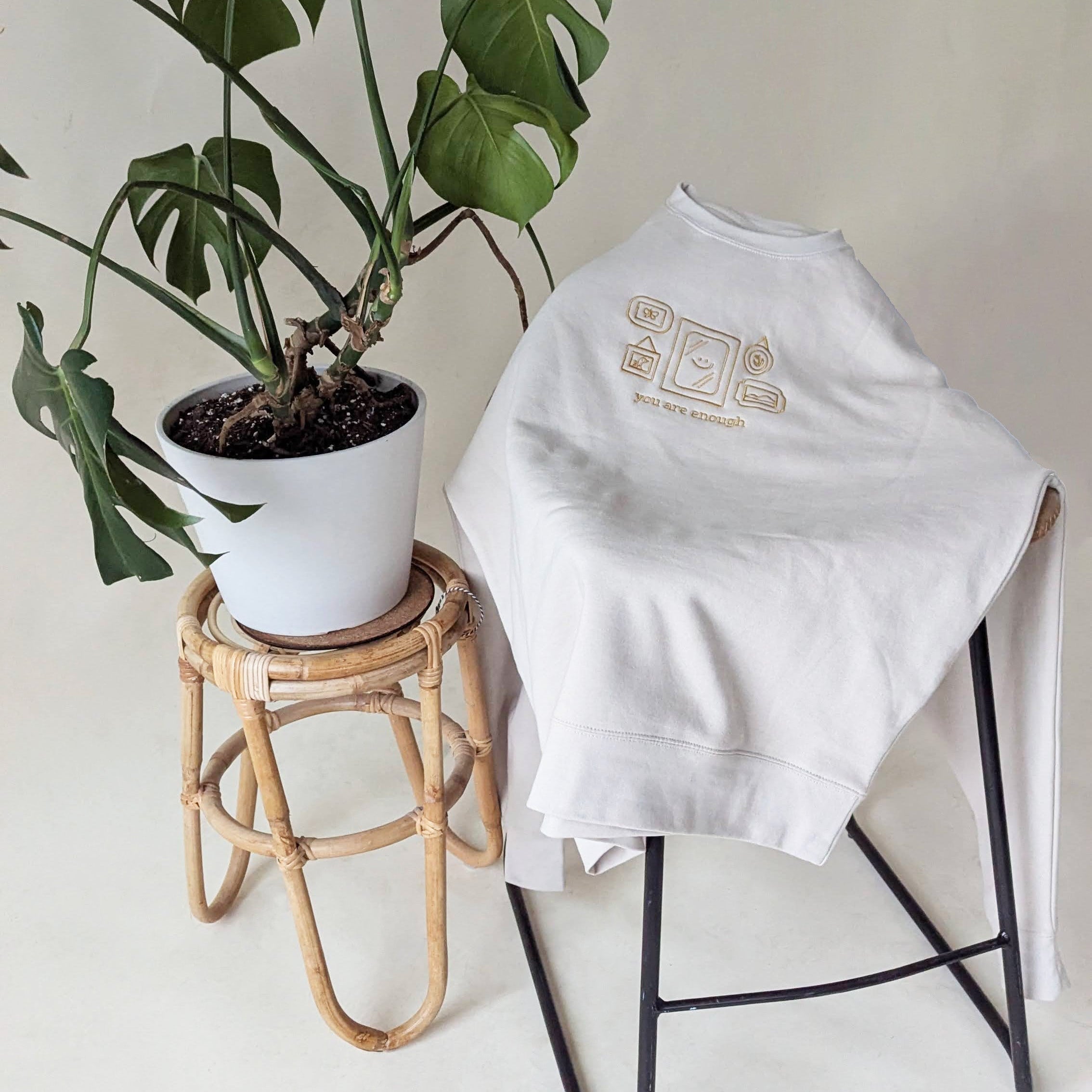 White sweatshirt with gold embroidery on a chair next to a potted monstera plant on a small stool. Sweatshirt features an illustration of a small gallery wall with a mirror in the center with a simple line drawing of a smiley face. Below the graphic is the text "you are enough".